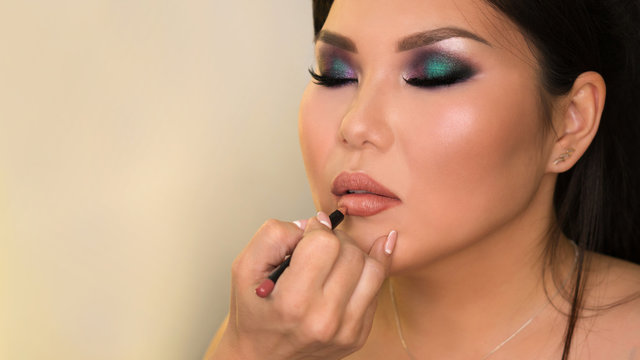 Makeup artist working with Asian model in beauty salon. MUA paints model's lips with contour pencil to make lips more expressive and seductive. Backstage. Photoshoot preparation. Beauty industry. Copy