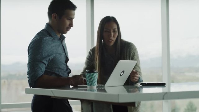 Tilt up to businessman and businesswoman talking and using laptop near window / Pleasant Grove, Utah, United States