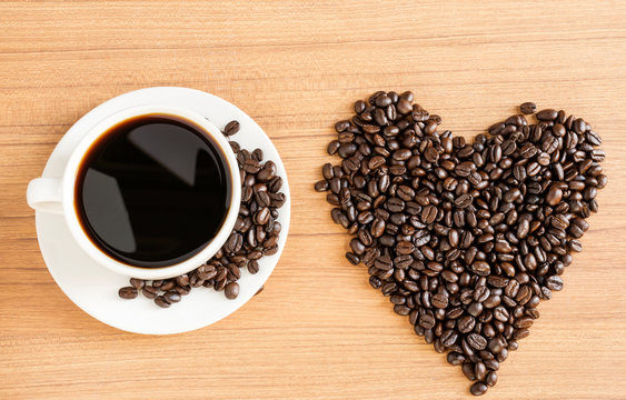 Fresh coffee has a fragrant aroma in a white cup. And coffee beans stacked into a heart shape on a wooden table top view - picture