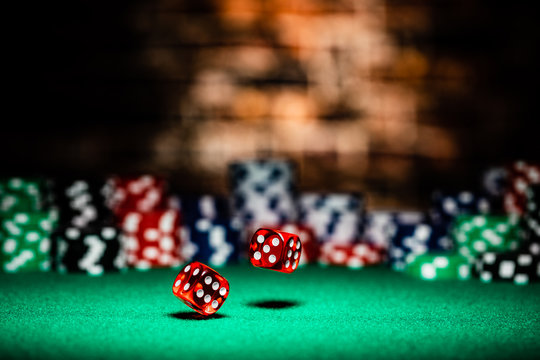 A very close up shot of 2 dice rolling on a green felt table top in very crisp focus, showing 4, 5 and 6 on the faces.  The background is a very soft focused wall of first chips, then a brick wall.