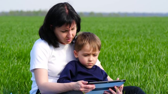 Mother and son relax in the Park sitting on the green grass. The family uses a tablet to play, watch a movie, or make a call. Technology, child development, freelancer.