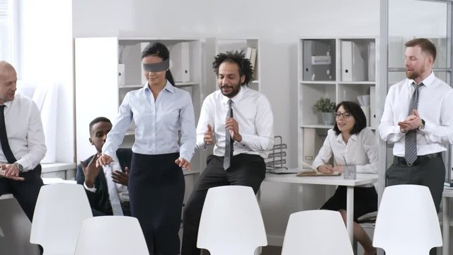 Medium shot of businesswoman in formalwear and blindfold walking between rows of chairs as her colleagues giving directions. Businesspeople playing trust game in office
