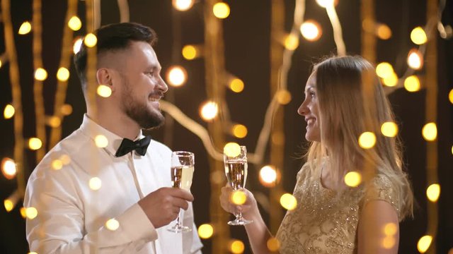 Medium shot of happy bearded man wearing shirt and bowtie and beautiful young woman in sparkly dress drinking champagne and chatting in room decorated with festive fairy lights