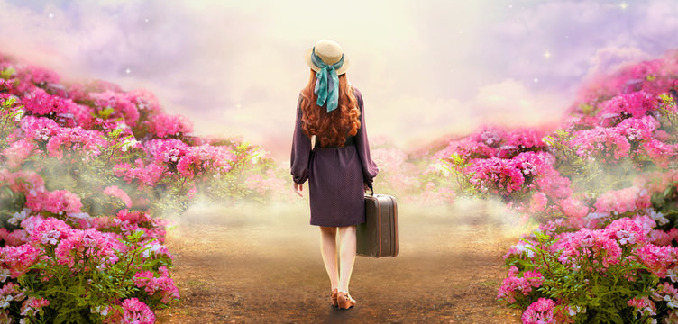 Young redhead woman in polka dot dress and hat with suitcase in retro style walking along a summer rose field path to mystical glow. Idyllic tranquil fantasy scene. Travel to fairytale concept.