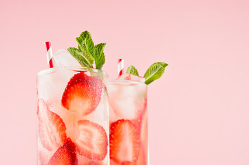 Homemade strawberry lemonades with slices, ice cubes, mint and straw in wet glasses on elegant pink color, closeup, top section, edge.