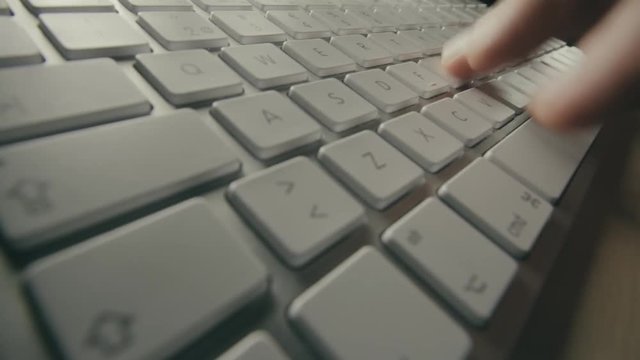 Close up view of silver computer keyboard with white keys and fingers typing. Person entering data into a computer. Fingers moving over keys and space bar. Dim light shines on metallic keyboard.