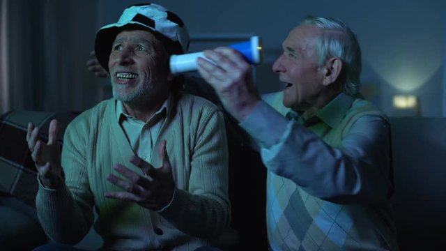 Senior football fans with hat and blowing horn hugging at home, leisure time