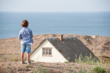 one lonely little kid in shirt and denim shorts standing on hill with old house and summer sea on...
