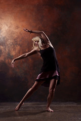 Full-length photo of young dancing blonde in short black dress on brown background