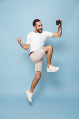Fototapeta na wymiar Full length photo of attractive caucasian man in casual white t-shirt screaming while jumping and taking selfie on smartphone