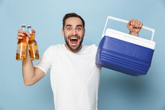 Photo of attractive caucasian man in casual white t-shirt laughing while carrying cooler box with beer bottles during summer party