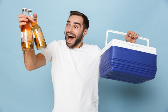 Photo of young caucasian man in casual white t-shirt laughing while carrying cooler box with beer bottles during summer party