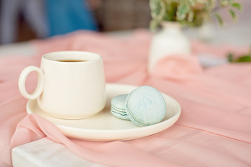 French blue macaroon and coffee cup standing on a wooden table with a pink tablecloth white vase with flowers roses and greens.