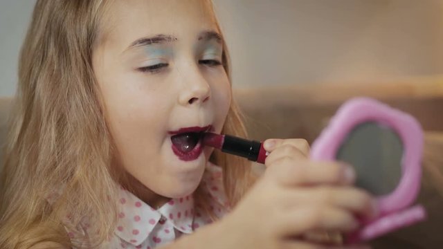 Portrait of a cute, blond schoolgirl who looks in a pink mirror and paints her lips with red lipstick, funny and careless makeup