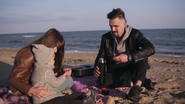 Young family in casualwear sitting on plaid by waterside and having hot drinks at picnic. Cold weather, weekend recreation