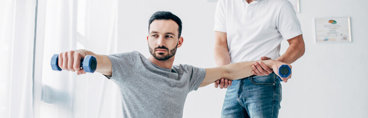 panoramic shot of chiropractor stretching arm of handsome patient with dumbbells in hospital