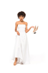 surprised african american bride touching wedding dress and holding shoes while standing isolated on white