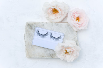 Fototapeta na wymiar Closeup of false eyelashes and beautiful flowers on the piece of marble stone on the white surface.Beauty concept