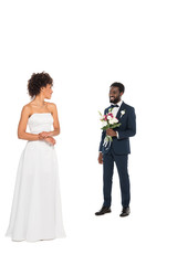 happy african american bride standing and looking at bearded bridegroom holding flowers isolated on white