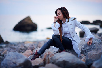 dreadlocks fashionable girl dressed in white jacket and black leather trousers posing near sea in...