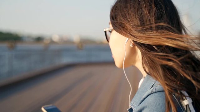 A young girl in glasses sits on the street and listens to music on headphones. The girl with the phone in her hands and in the headphones, her hair fluttering in the wind. Slow motion.