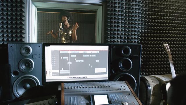 Tilt down of black male sound engineer in headphones sitting in control room and using mixing desk as young African-American woman in sparkly dress singing into microphone in recording studio