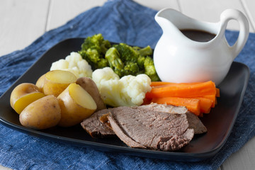Lamb roast Sunday lunch dinner with vegetables on a aquare black plate, with gravy jug, on a blue tea towel.  White wood background
