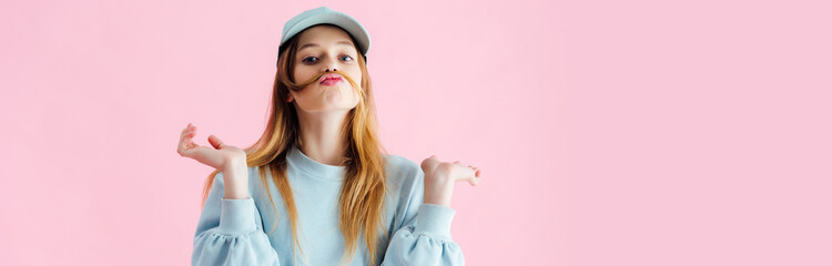 panoramic shot of pretty teenage girl in cap with mustache of hair grimacing isolated on pink