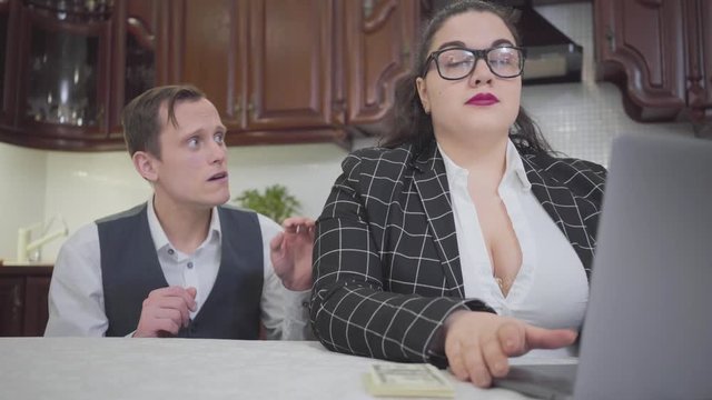 Young confident plump woman in glasses at the table in kitchen working with laptop. A timid young man asking more money and she gives him dollars. Family relationship.