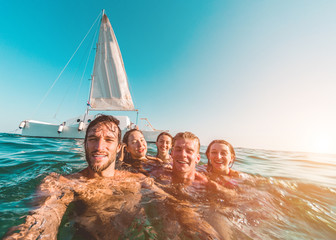 Happy friends taking a selfie with action cam inside the ocean with sail boat in background - Young...