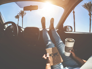 Woman drinking coffee paper cup inside car with feet on dashboard - Girl relaxing in auto trip reading traveler book with ocean beach and palms in background - Travel concept - Focus on hands