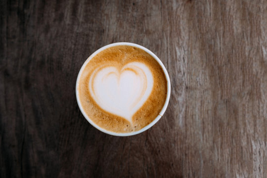 Paper cup of cappuccino coffee with beautiful latte art in the shape of heart on wooden background. Top view, copy space. 