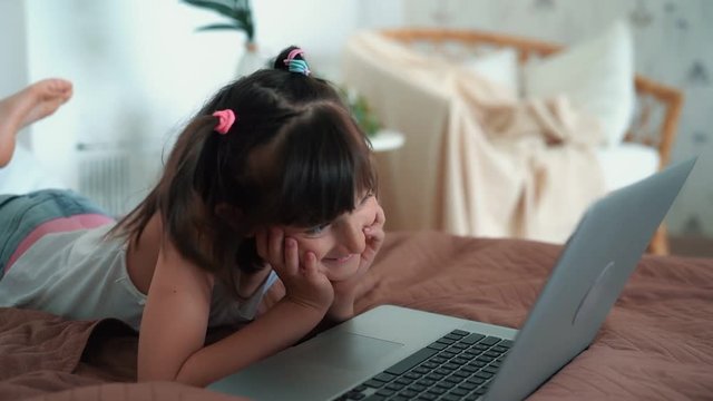 Little girl lying on bed, uses laptop, plays computer game, slow motion