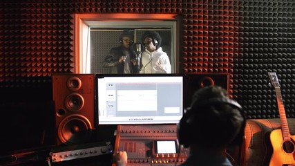 Dolly-in shot of black man and woman dancing and singing into microphone in isolation booth of recording studio as male sound engineer using mixing desk in control room - Powered by Adobe