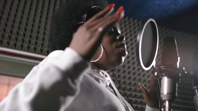 Handheld low angle shot of beautiful black female singer in headphones and hoodie singing soulful song into microphone in live room of recording studio