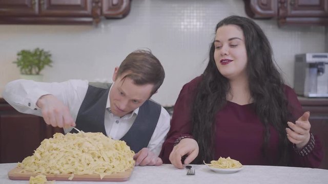 Portrait of pretty confident pretty plump woman and skinny man in the kitchen at the table in front of a big and small plate s of noodles. Funny family.