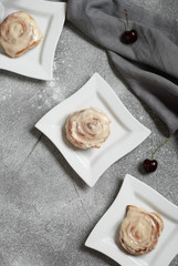 Cinnamon buns in square plates on a gray background, decorated with a linen towel and cherry. Vertical image. Copy space. Flat lay.