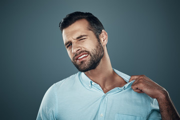 Uncomfortable. Tired young man pulling collar while standing against grey background