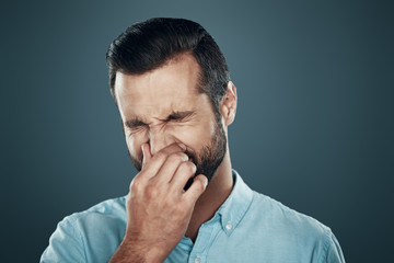 Allergy. Handsome young man covering nose with hand while standing against grey background