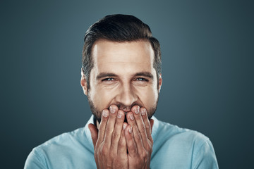 Sincere fun. Handsome young man laughing and covering mouth with hands while standing against grey background