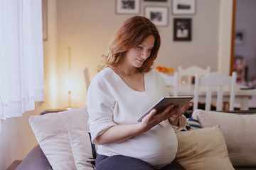 Beautiful Caucasian pregnant woman sitting in living room in sofa and using tablet.