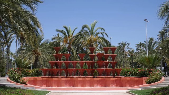 An interesting fountain with many goblets flowing down the water. In the background many palm trees in the Spanish city Elche at the Palms Park. Slow Motion 4K.