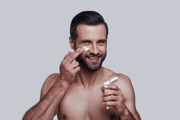 Fototapeta premium Beauty treatment. Handsome young man applying moisturizer and smiling while standing against grey background