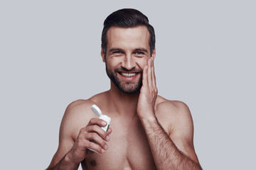 Fototapeta premium Personal care. Handsome young man applying moisturizer and smiling while standing against grey background