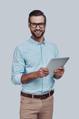 Ready to help. Handsome young man using digital tablet and smiling while standing against grey...