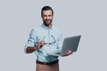 May I help you? Good looking young man carrying laptop and looking at camera while standing against grey background