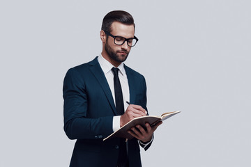 Making notes. Handsome young man writing something down while standing against grey background