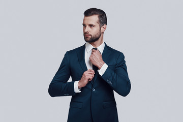 Getting ready. Handsome young man adjusting his necktie while standing against grey background