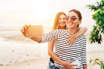 Two caucasian girlfriends hugging and posing for selfie on the sea side using smartphone. Careless...
