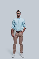 Confident businessman. Full length of good looking young man carrying laptop and looking at camera while standing against grey background